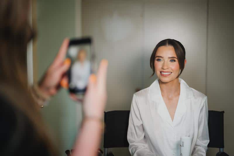 Woman in white blouse smiling while being photographed with a smartphone indoors.