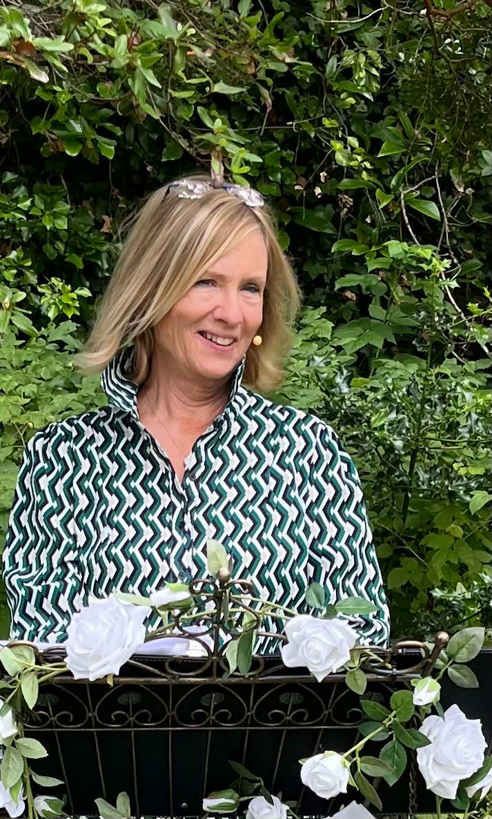 Woman smiling in a garden setting, wearing a patterned blouse, standing behind a floral-decorated railing.