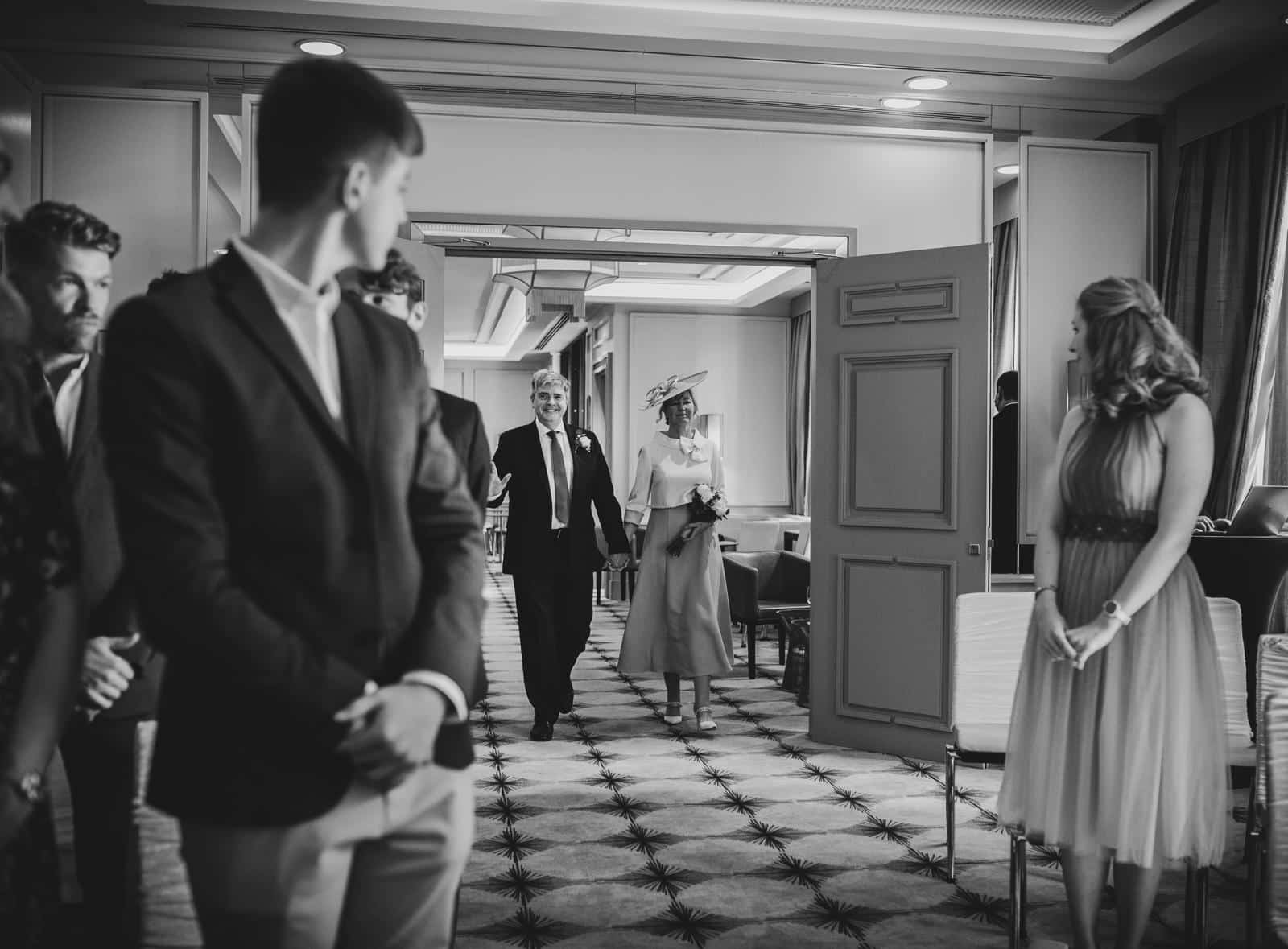 A wedding couple walks through a doorway as guests look on, in a formal indoor setting. Black and white image.