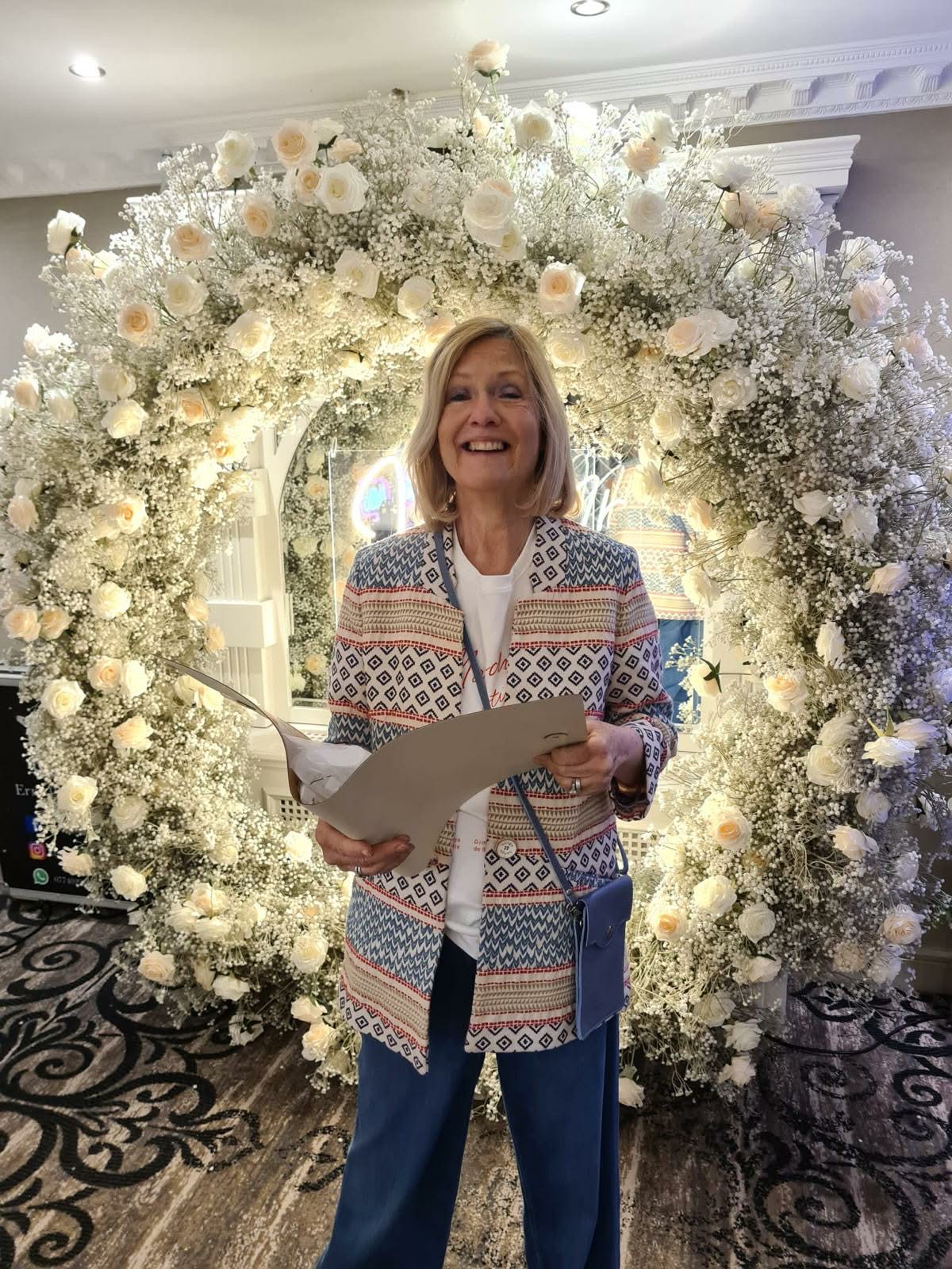 Smiling woman holding papers, standing in front of a large circular floral arrangement with white and peach roses.