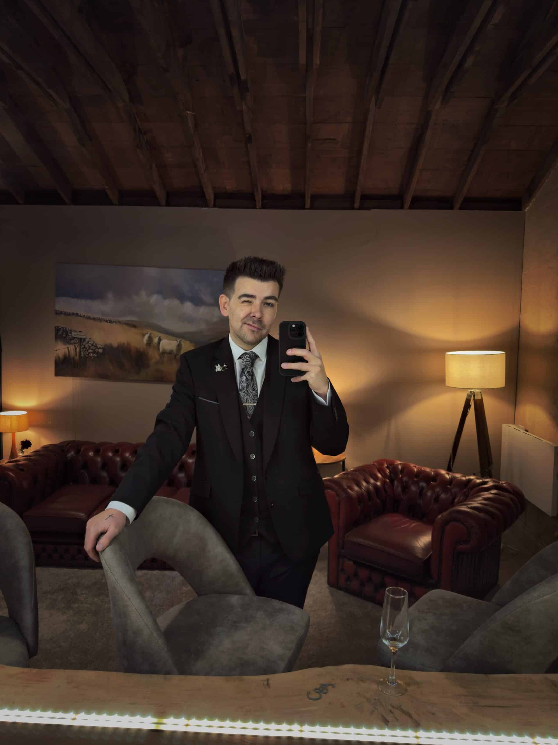 Man in a suit taking a selfie in a cozy room with leather sofas and a wine glass on the table.