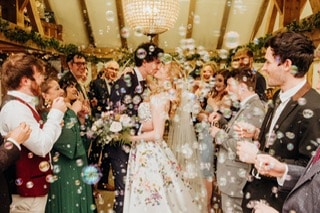 Couple kissing at a wedding surrounded by guests and bubbles.