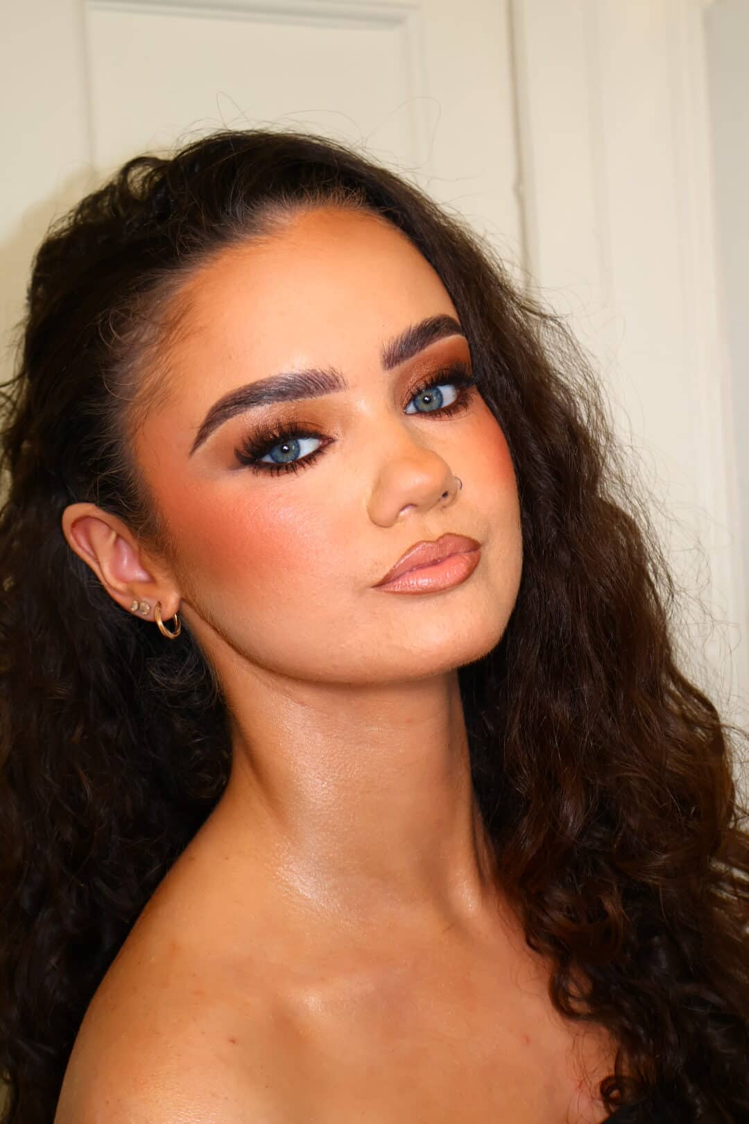 Woman with curly hair and vibrant makeup, featuring blue eyes, prominent eyebrows, and peachy lipstick.