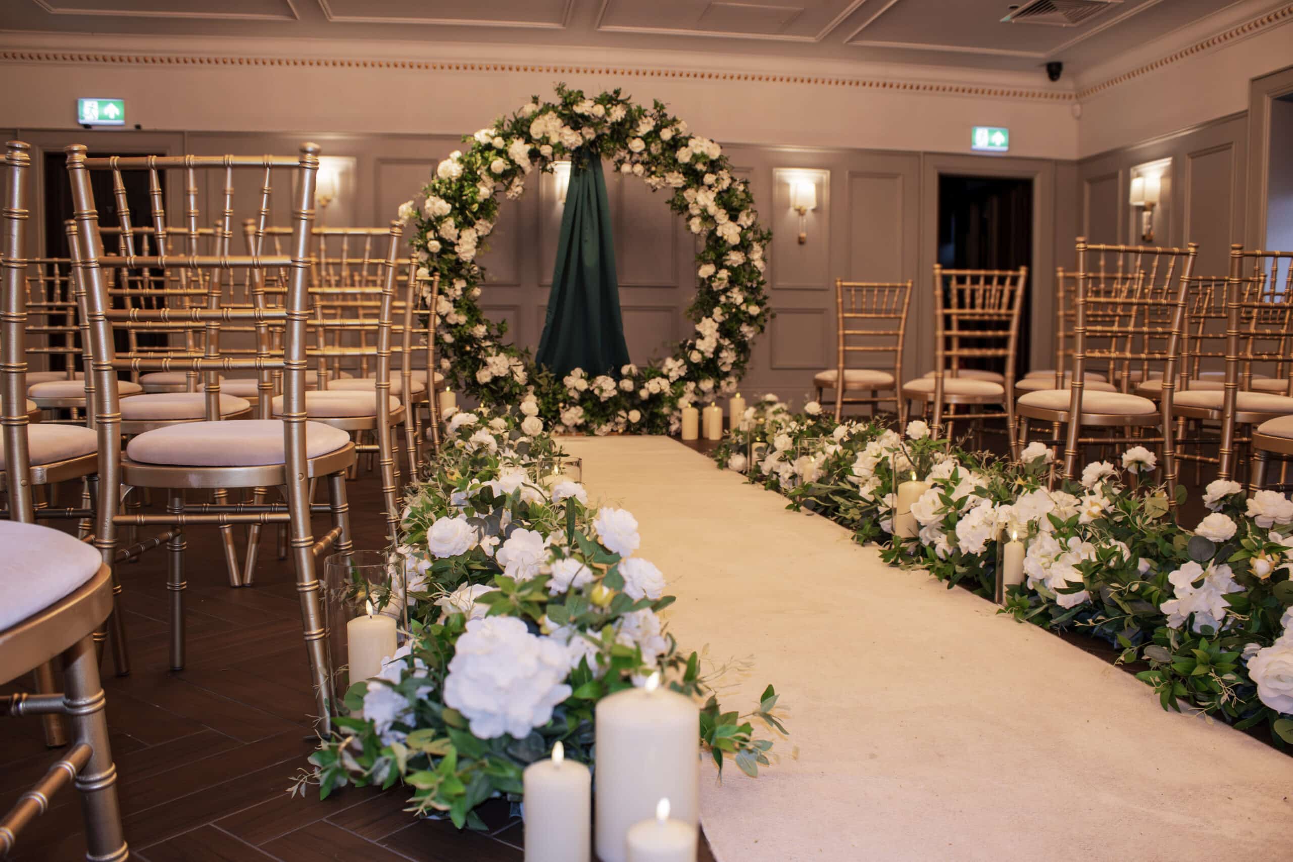 Elegant indoor wedding aisle with floral arch, gold chairs, white flowers, and candles.