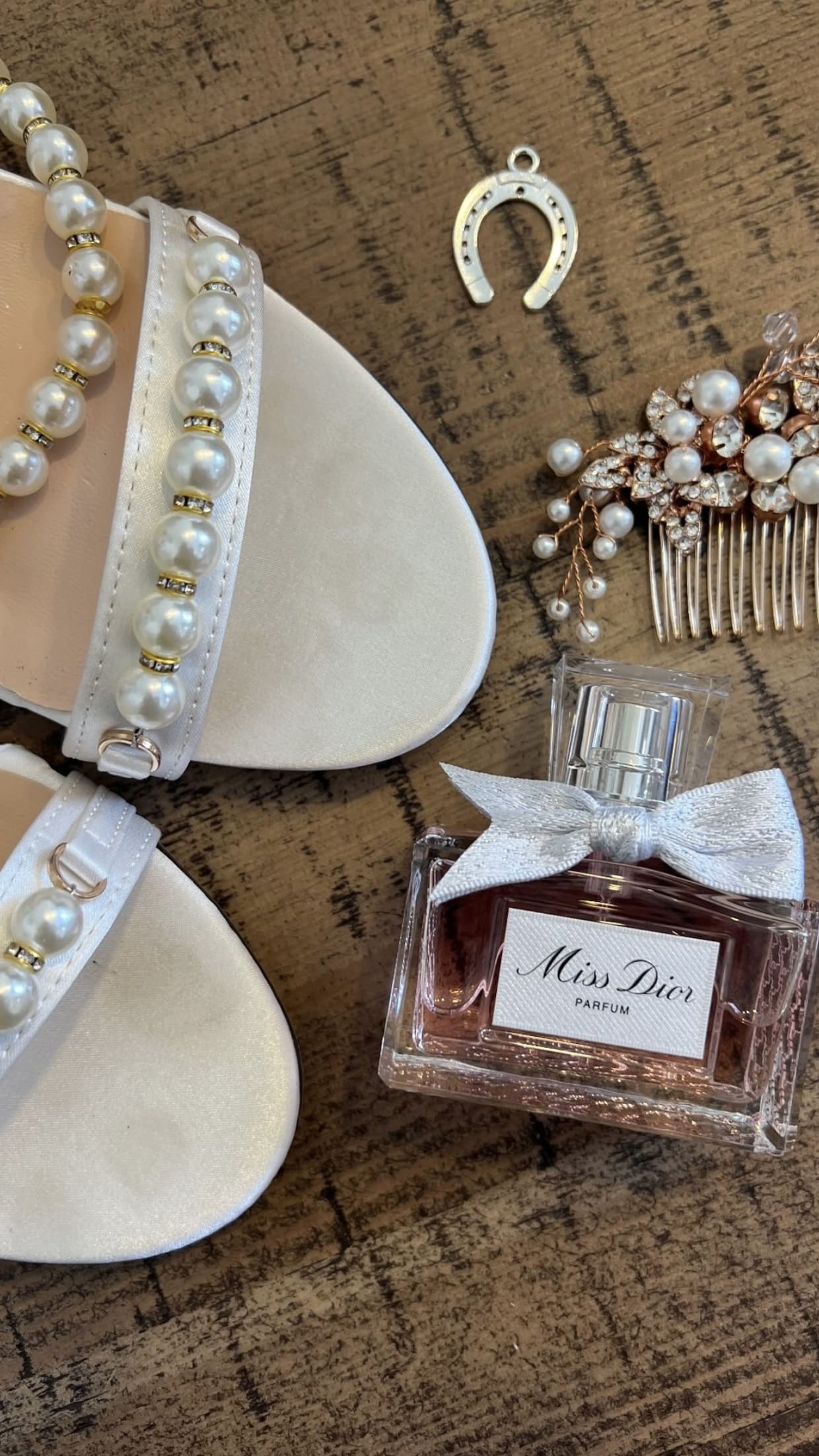 Pearl sandals, Miss Dior perfume, a horseshoe charm, and pearl hair accessories on a wooden surface.