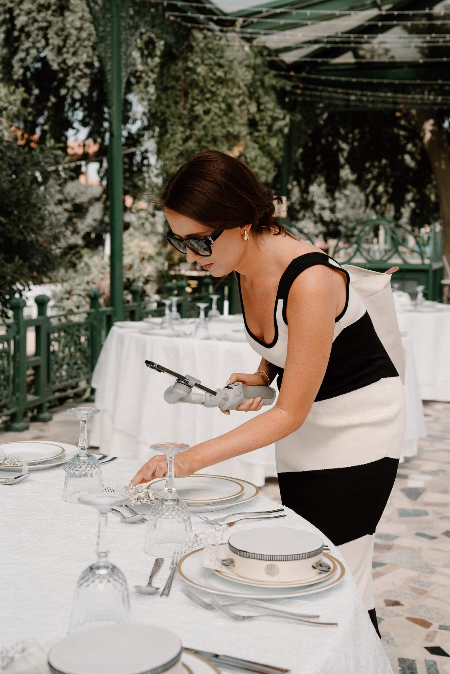 A woman setting up an elegantly arranged table outdoors, holding a camera stabilizer.