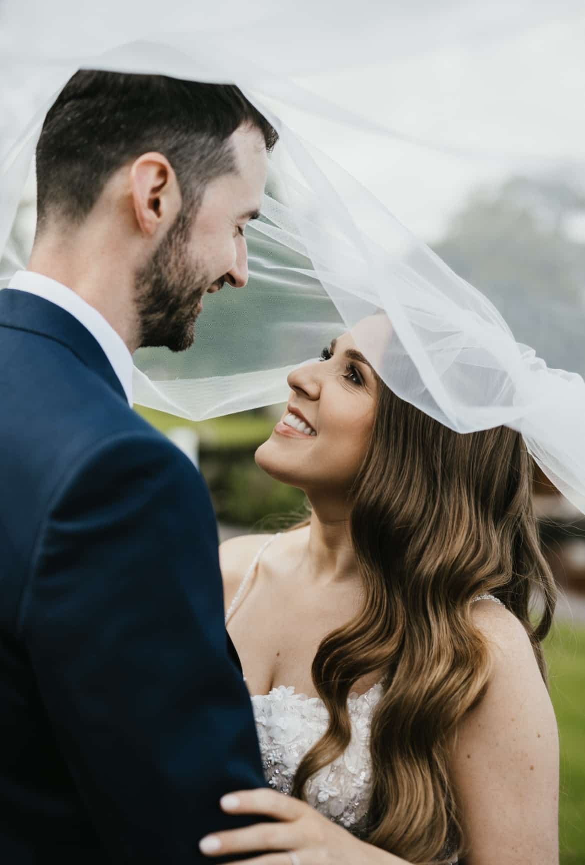 Bride and groom smiling under a white veil on their wedding day.