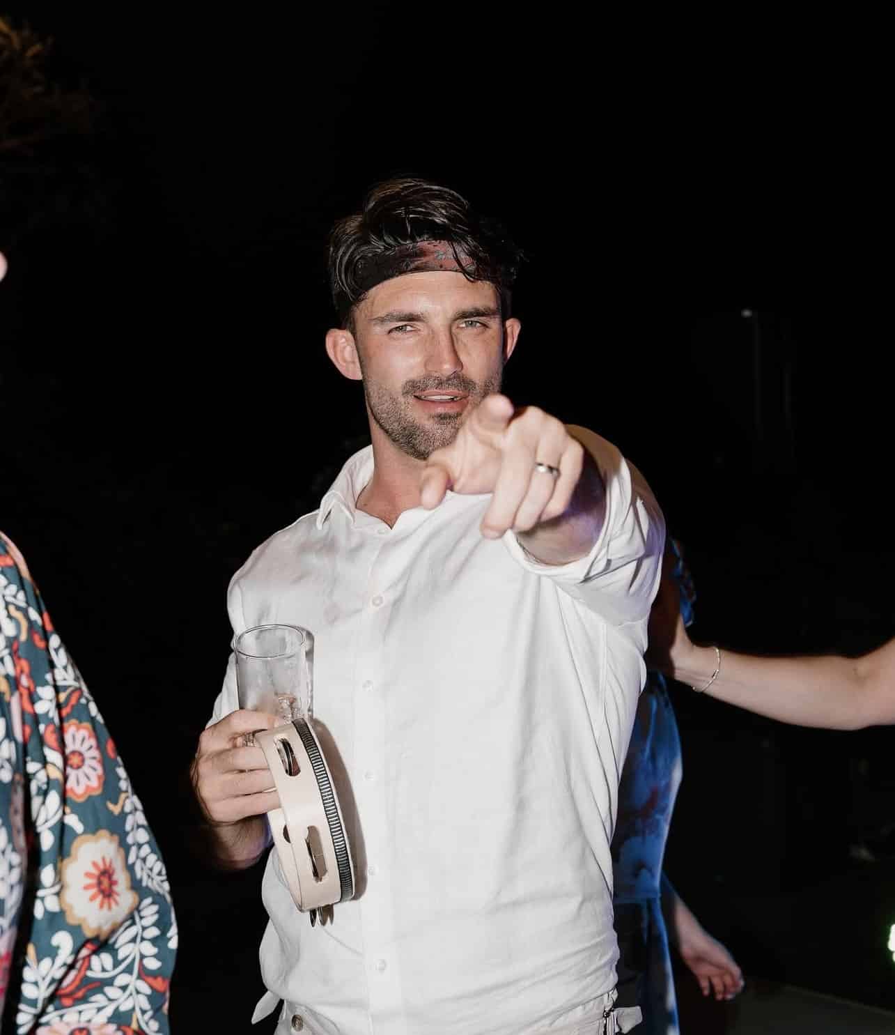 Man in a white shirt holding a tambourine and drink, pointing at the camera in a party setting.