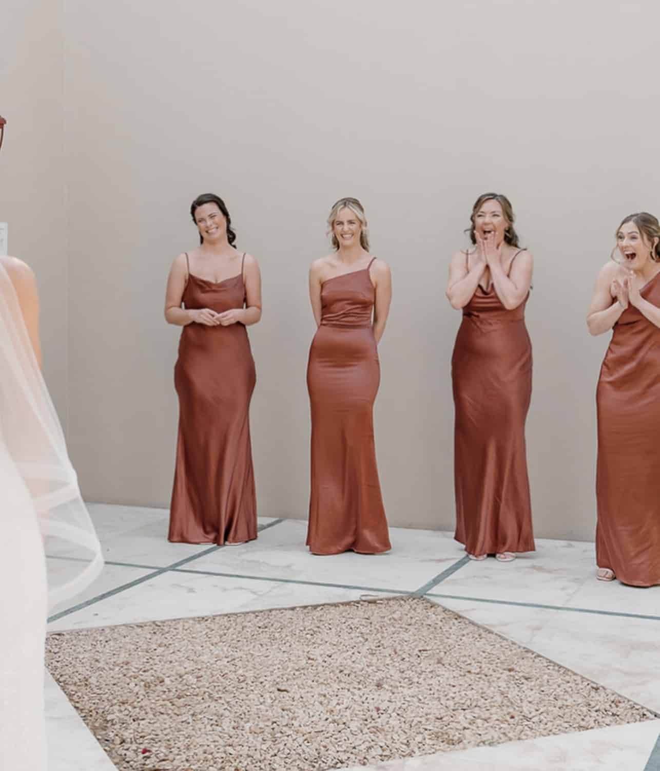 Bridesmaids in bronze dresses react with excitement and joy, facing a bride in a white gown and veil.