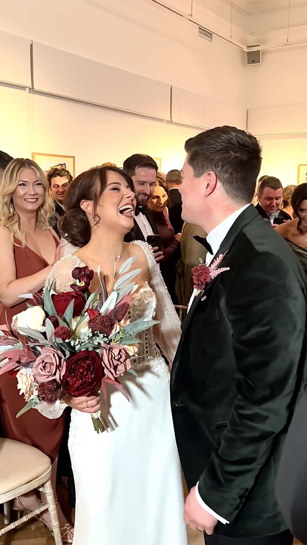 Bride and groom laughing joyfully at their wedding, surrounded by guests. Bride is holding a bouquet of red and pink flowers.