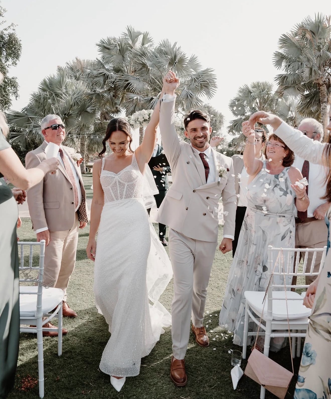 A bride and groom celebrate while walking down the aisle outdoors, surrounded by guests and palm trees.