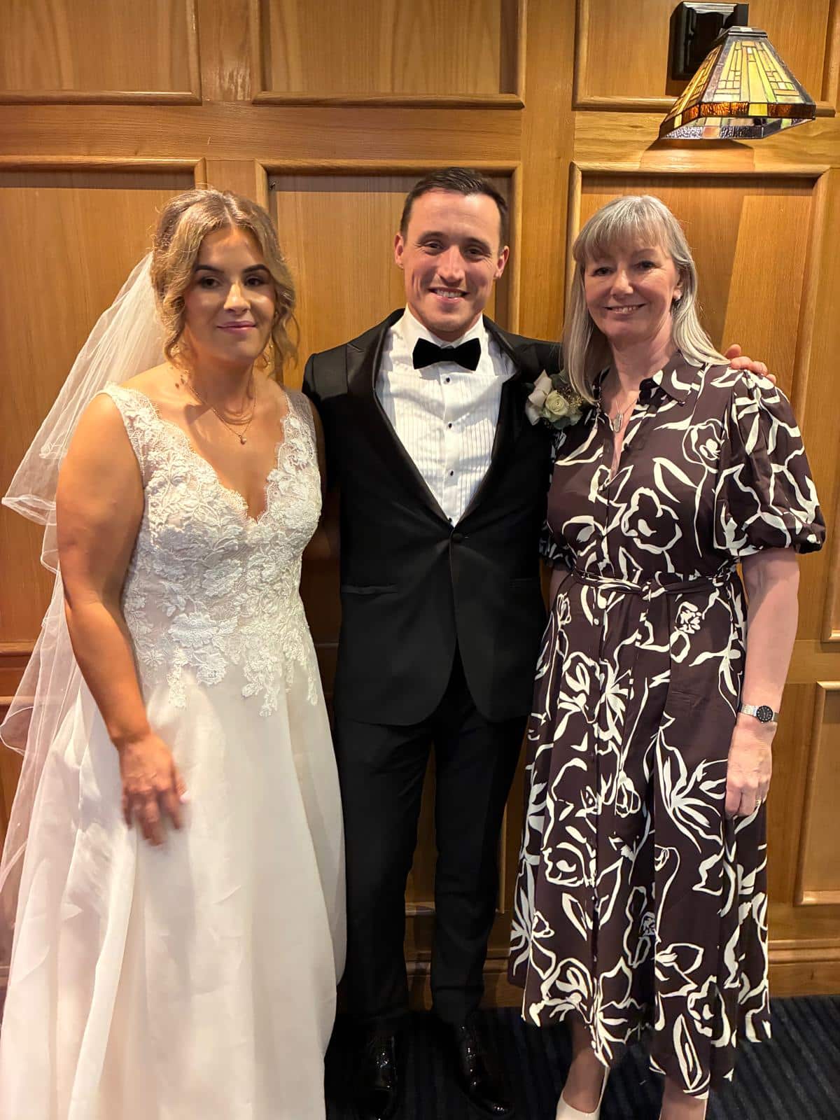 Bride and groom in wedding attire with a guest in a floral dress standing indoors.