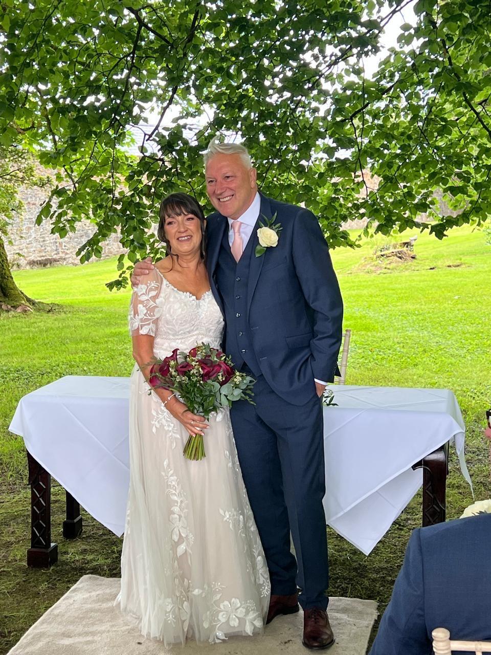 Bride and groom smiling under a tree canopy, with the bride holding a bouquet of red flowers.