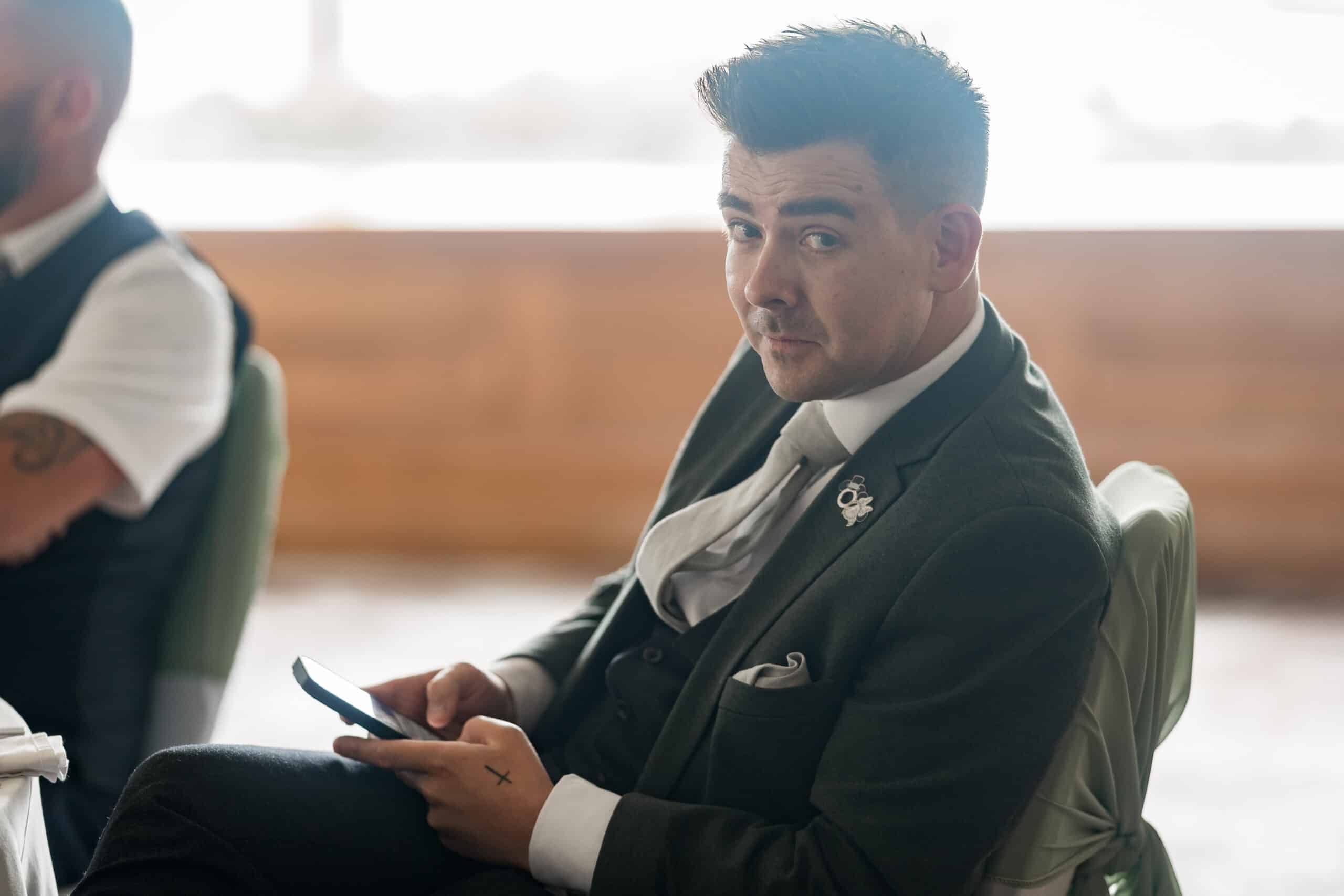 Man in a suit sitting at a table, holding a smartphone and looking at the camera.