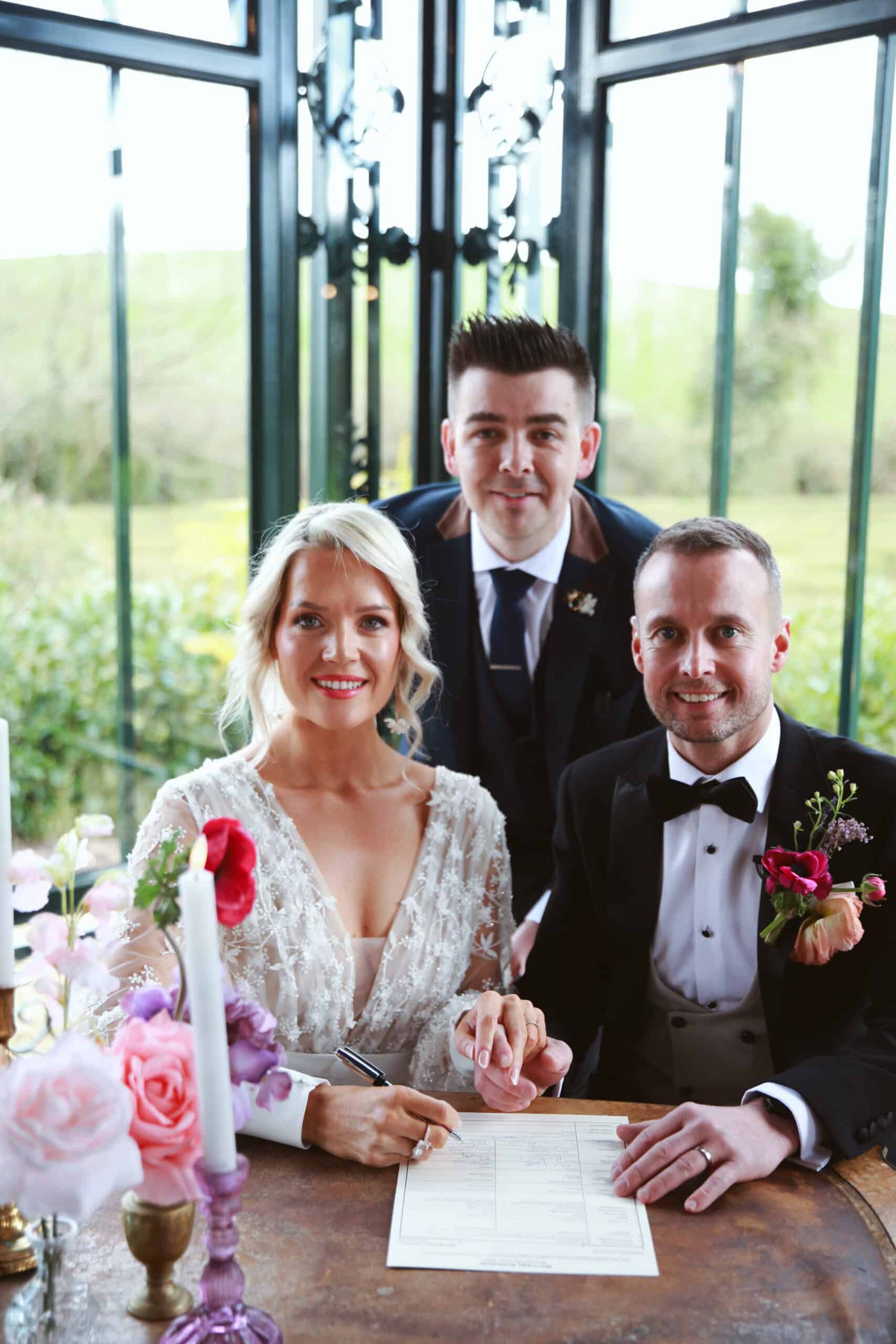 Bride and groom signing a marriage certificate with a man in a suit standing behind them.