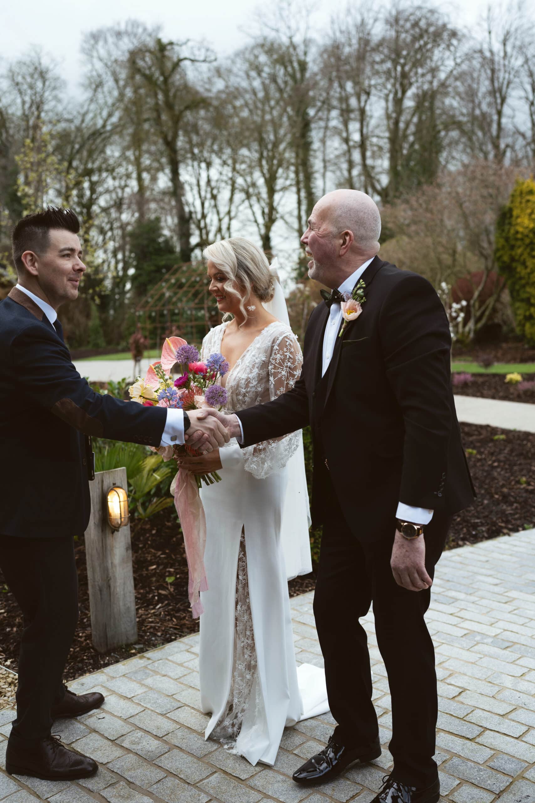 Groom shaking hands with a man, bride holding colorful bouquet, standing outdoors on a cloudy day.
