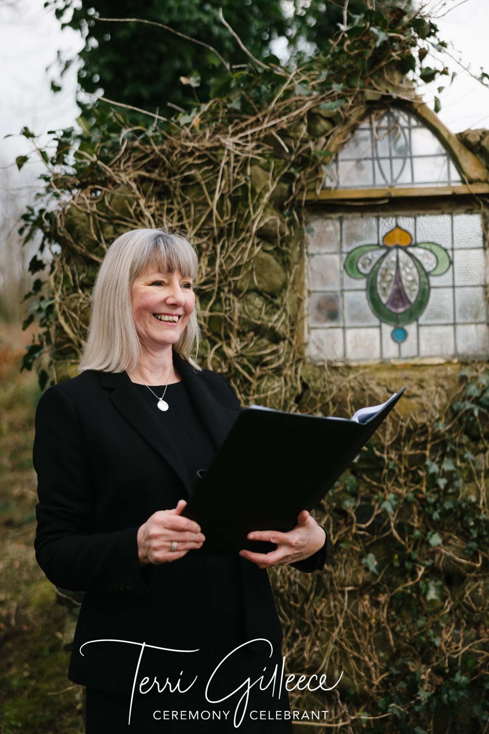 Smiling woman holding a black folder in front of a decorative window with ivy.