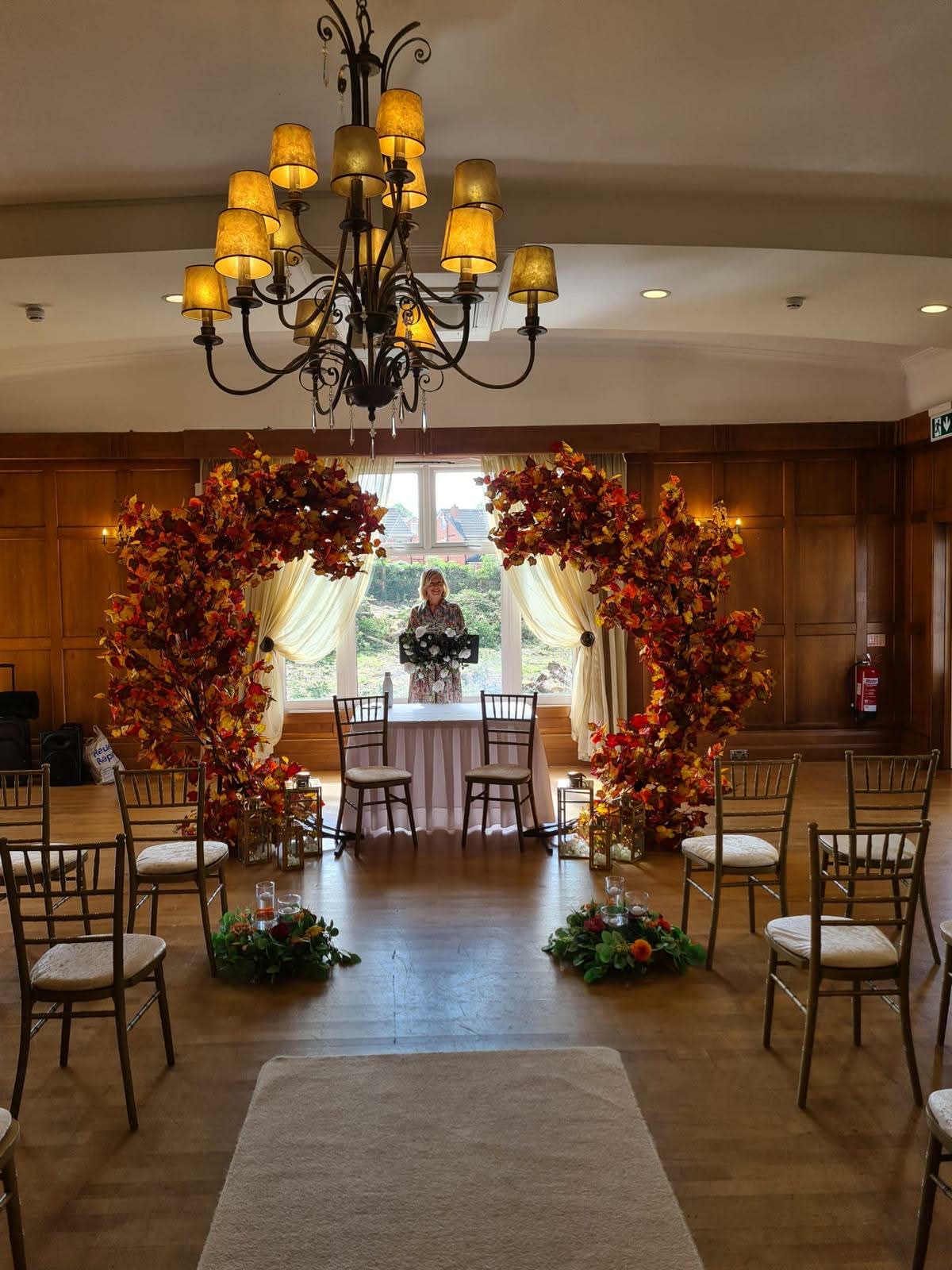 Indoor wedding ceremony setup with fall-themed floral arches, chairs, and a chandelier.