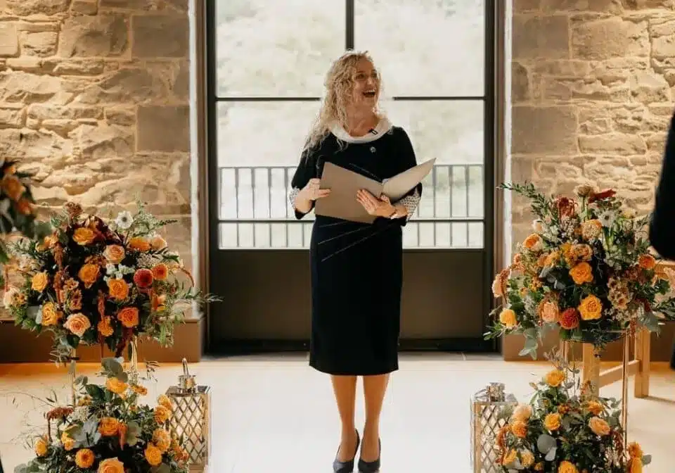 Celebrant leading a ceremony indoors with floral arrangements, featured in vendor listing card.